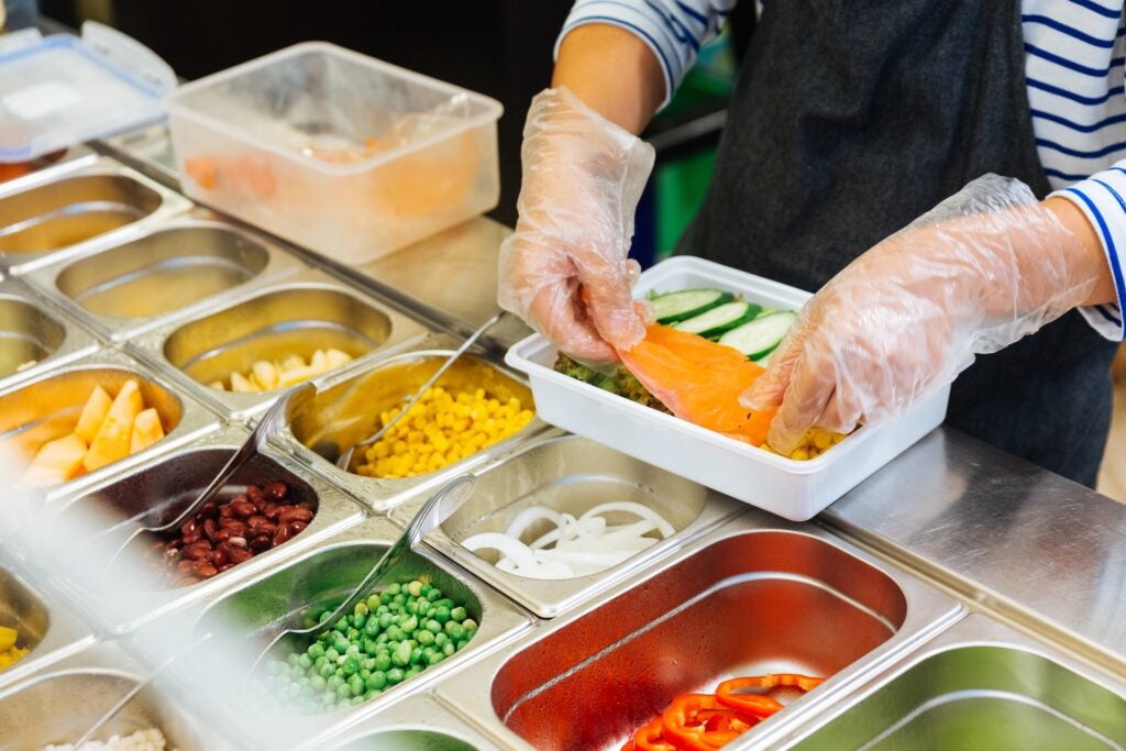 close up of someone wearing gloves and an apron putting a dish together on the line with food prep containers