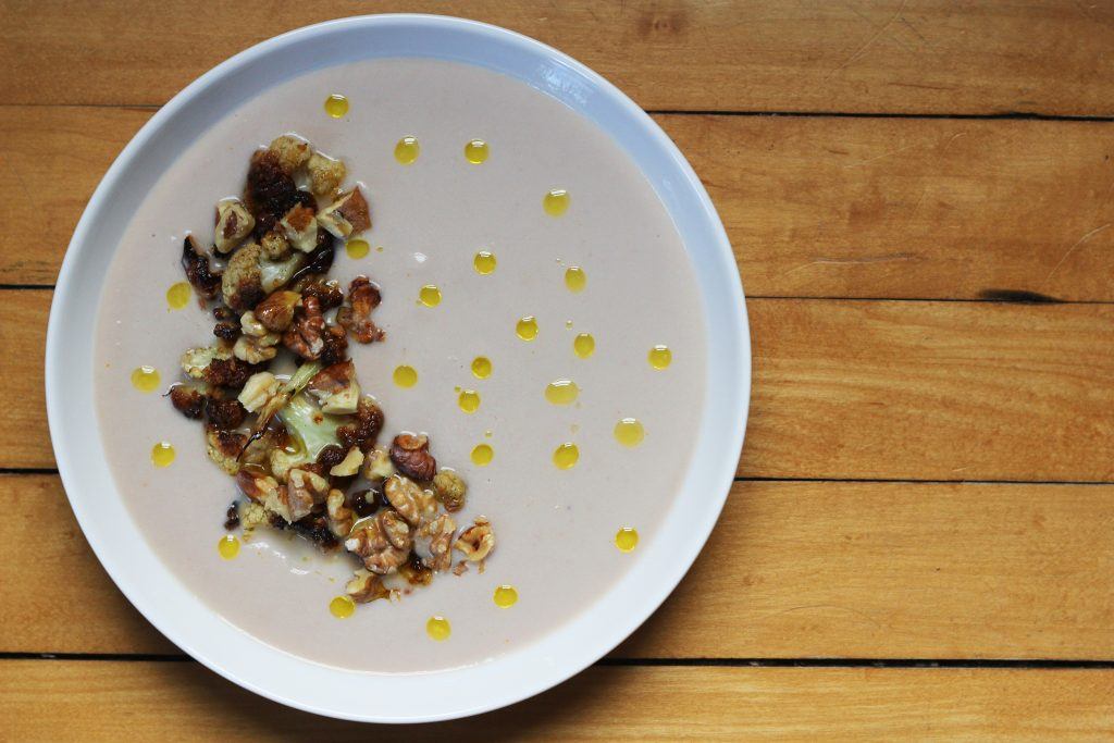 A bowl of creamy cauliflower soup topped with roasted cauliflower and toasted walnuts and drips of extra virgin olive oil